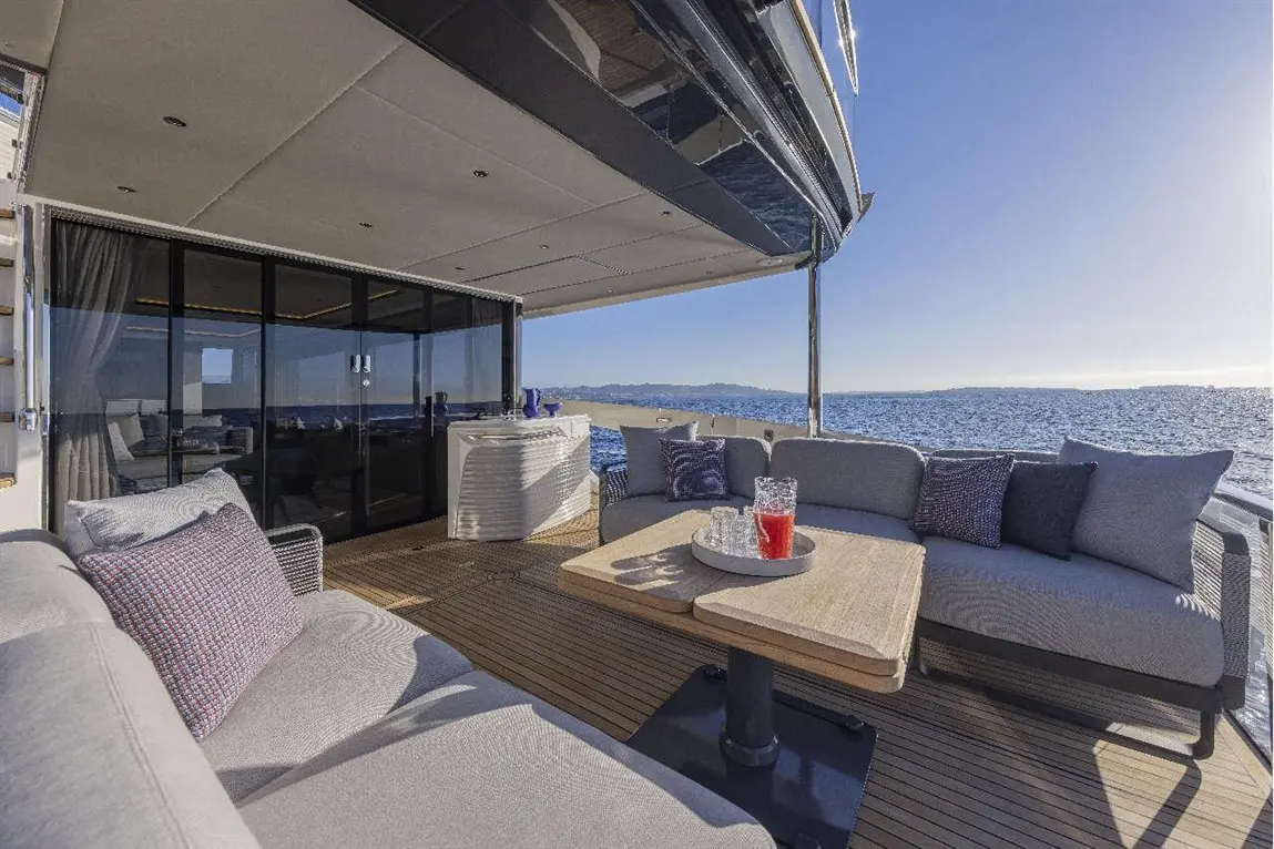 Luxury yacht deck with plush seating, a wooden table, and drinks, overlooking a serene sea and sunny sky.